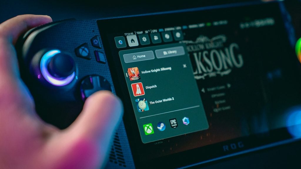 A close-up of a handheld gaming console screen displaying a game library, with a user's hand holding the device and colorful lights reflecting on the console.