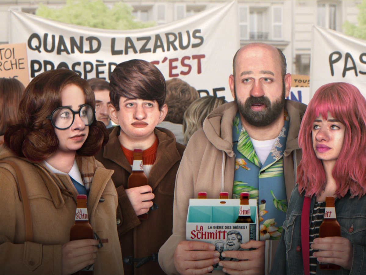 Four people holding beer bottles stand in front of a crowd with protest signs written in French. One person holds a beer six-pack. The background appears to be an outdoor public space.