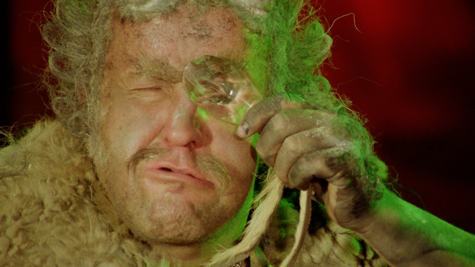 A person with messy hair and a fur garment holds a large crystal to their eye, squinting, with green light illuminating their face.