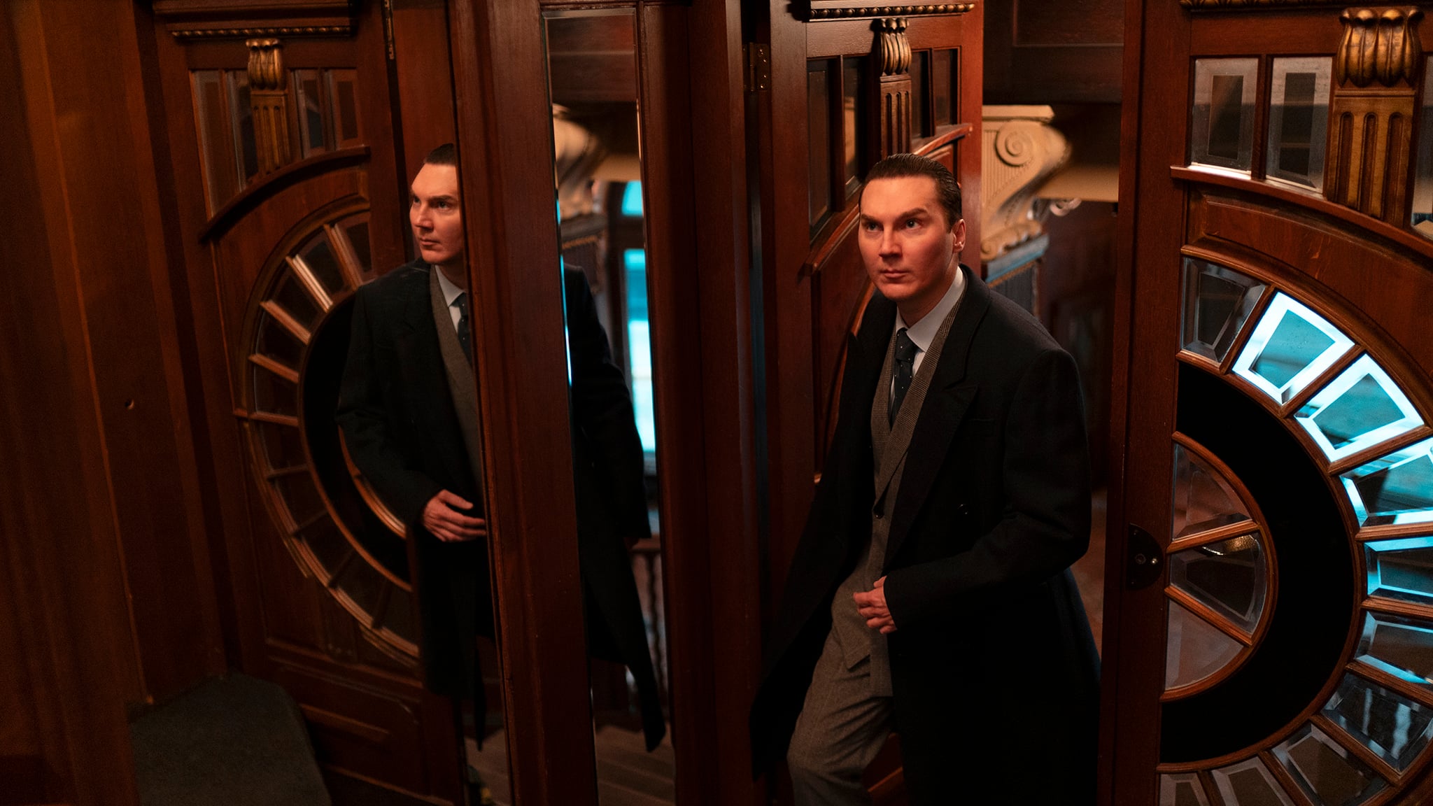 A man in a suit and overcoat stands by a wooden door with glass panels, reflected in a mirror, with ornate woodwork and circular window details in the background.
