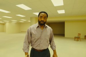 A man with a beard stands in a large, empty room with bright ceiling lights and beige walls. There are a few wooden chairs against the walls in the background.