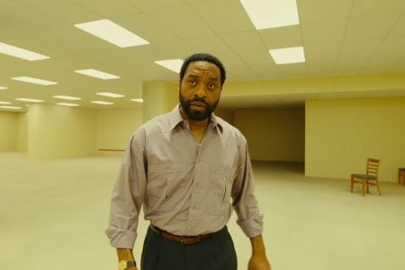 A24 To Release The Creepy Box Office Horror Hit ‘Backrooms’ On 4K UHD Blu-Ray This August A man with a beard stands in a large, empty room with bright ceiling lights and beige walls. There are a few wooden chairs against the walls in the background.