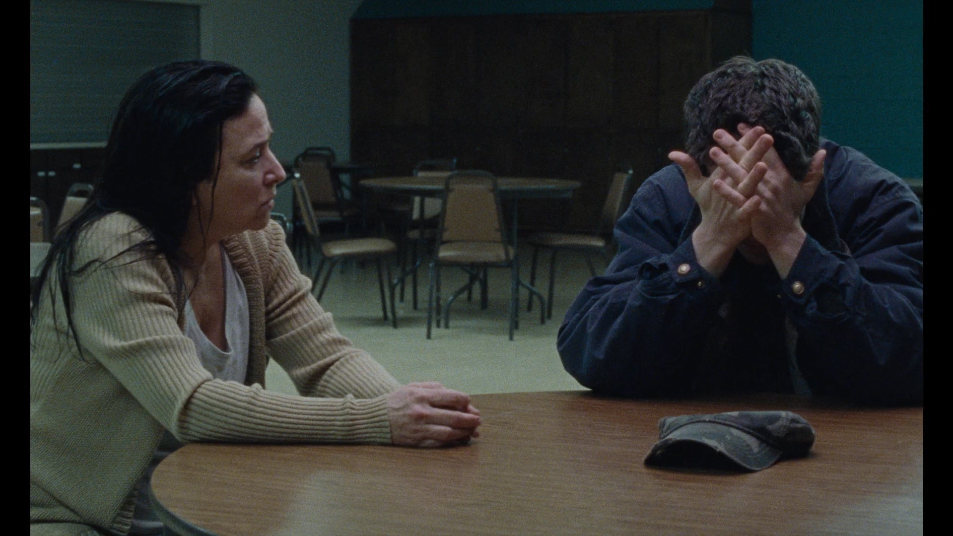A woman sits at a round table facing a man who covers his face with his hands, while a cap lies on the table between them in a sparse room with empty chairs.