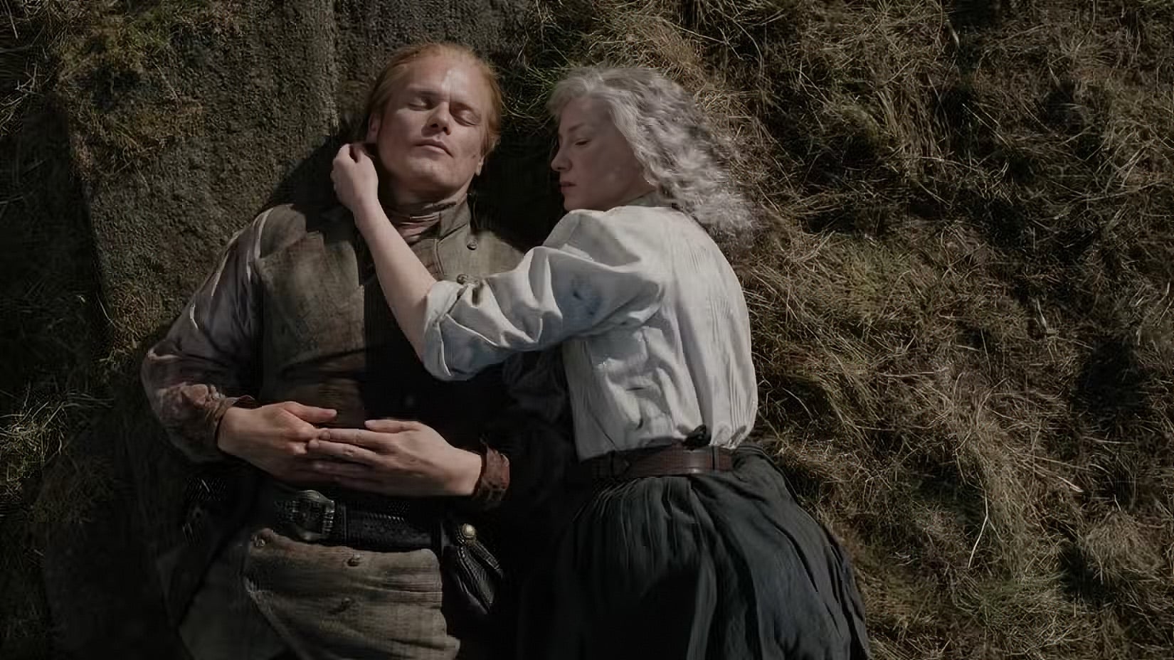 A man and a woman lie on the grass, the woman resting her head on the man's chest. Both appear calm and are dressed in historical clothing.