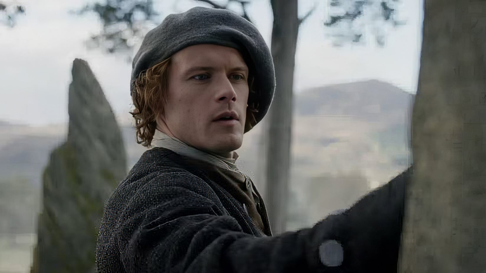 A man wearing historical clothing and a beret stands outdoors, touching a large stone, with trees and hills visible in the background.