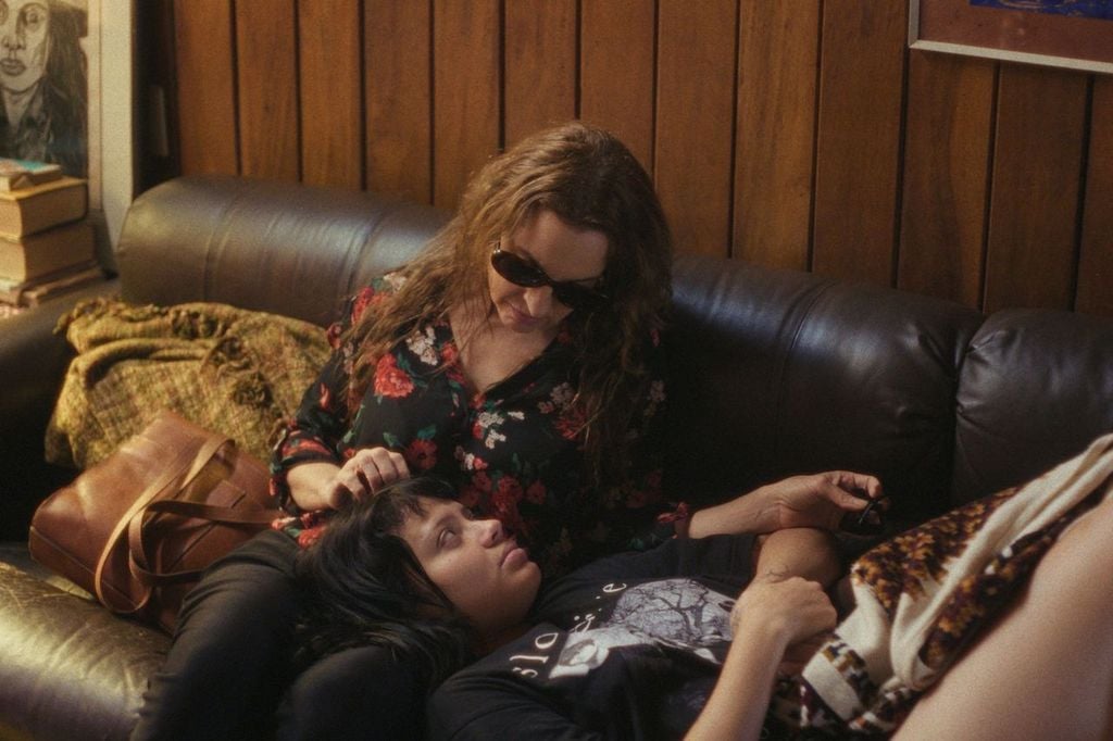 Two women are on a dark couch; one wears sunglasses and sits upright, while the other lies with her head in the first woman's lap. Wooden panel wall and a stack of books are visible in the background.