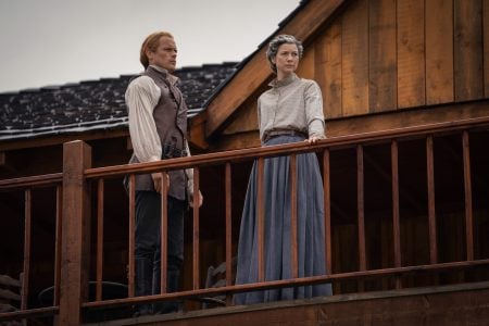 Two people in period clothing stand on a wooden balcony, looking into the distance; a rustic wooden building serves as the backdrop.