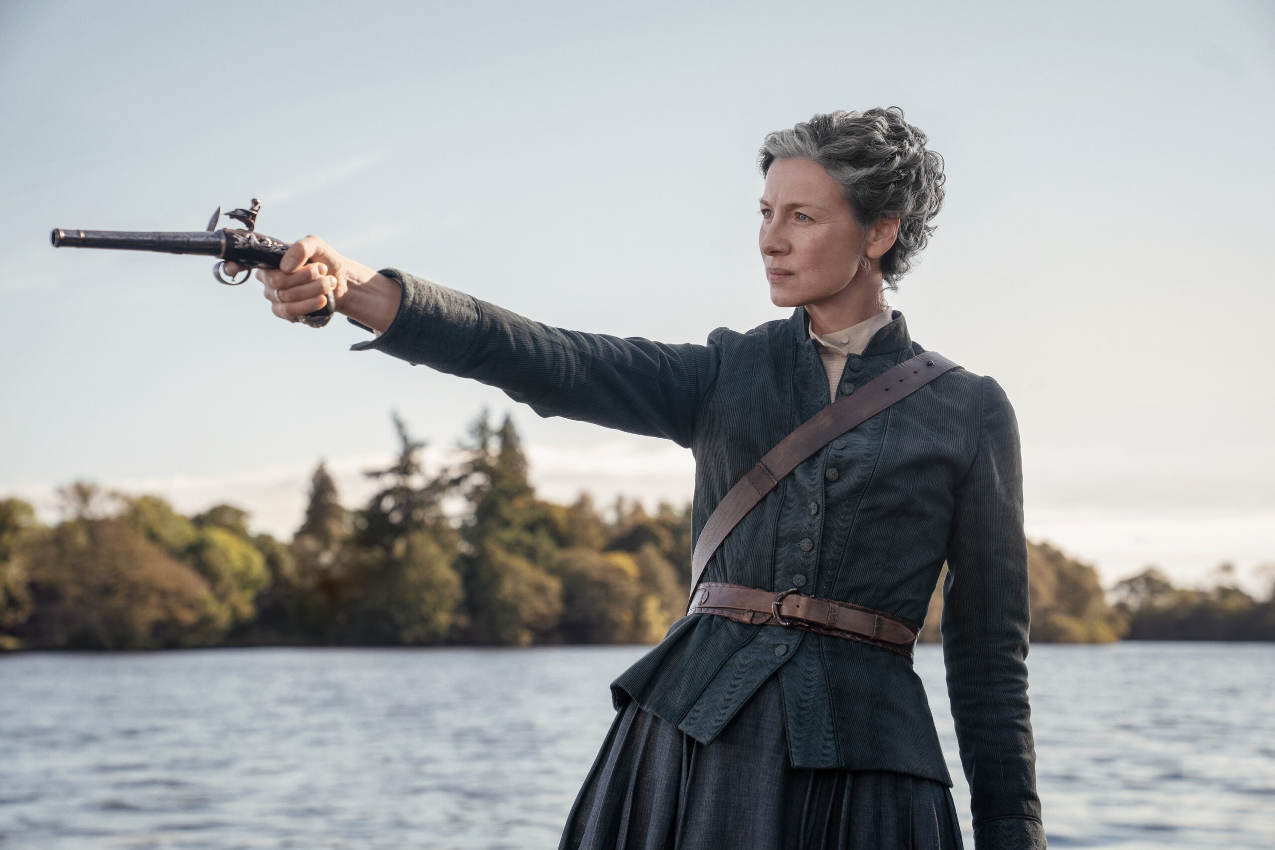 A woman in historical clothing stands by a lake, aiming a flintlock pistol with her right arm extended. Trees and blue sky are visible in the background.