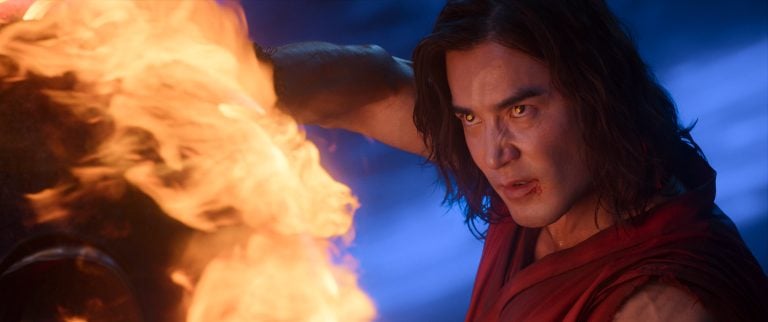 A man in a red outfit intensely holds his arm near large flames, his face illuminated by the fire and set in concentration against a dark, blue-lit background.