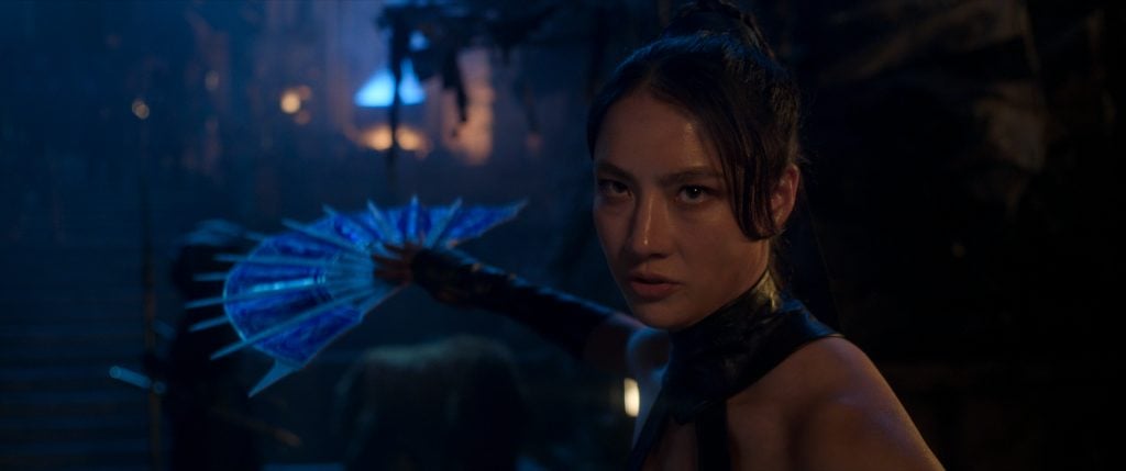 A woman in a dark setting holds an open, blue-bladed fan in a defensive stance, looking intently forward.