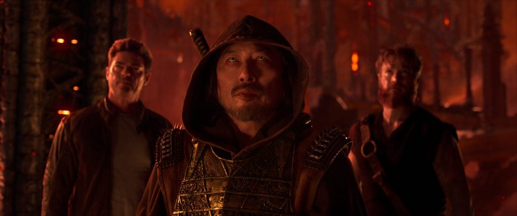 Three men stand together in a dimly lit, fiery setting; the central figure wears a hooded, armored outfit, while the other two appear in rugged clothing.