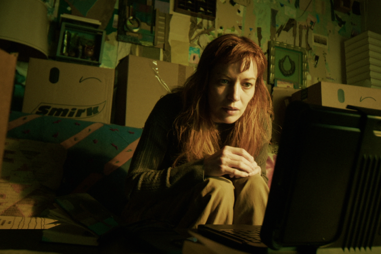 A woman with long red hair sits on the floor surrounded by cardboard boxes, looking intently at a laptop in a dimly lit room.