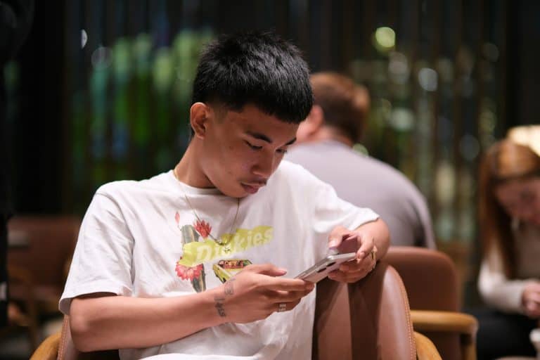 A young man in a white T-shirt sits on a chair, looking at his smartphone, with blurred people and a dark background behind him.