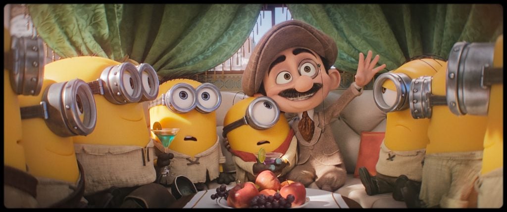 A group of four Minions sit on a couch with a man in a hat and suit, surrounded by fruit, with curtains in the background.