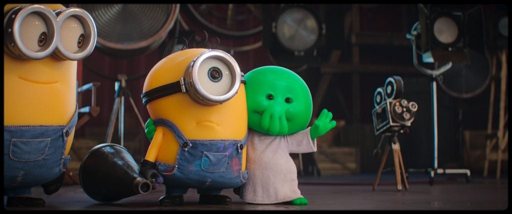 A yellow Minion in overalls stands next to a small, green, tentacled character wearing a white shirt, with film equipment visible in the background.