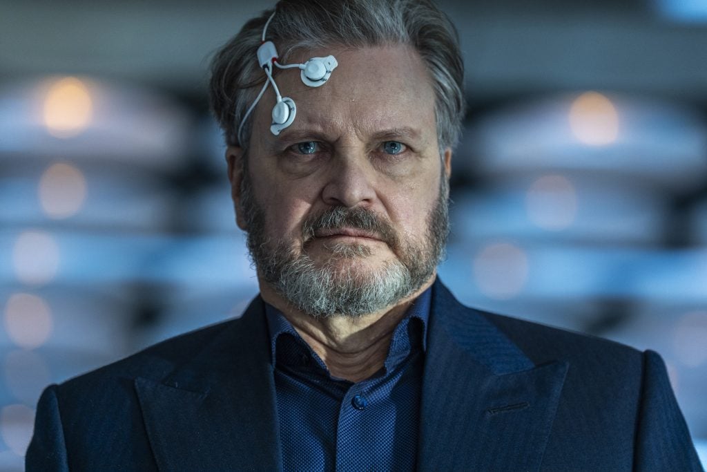A man with gray hair and a beard wears a blue suit and has a medical or technological device attached to his forehead.