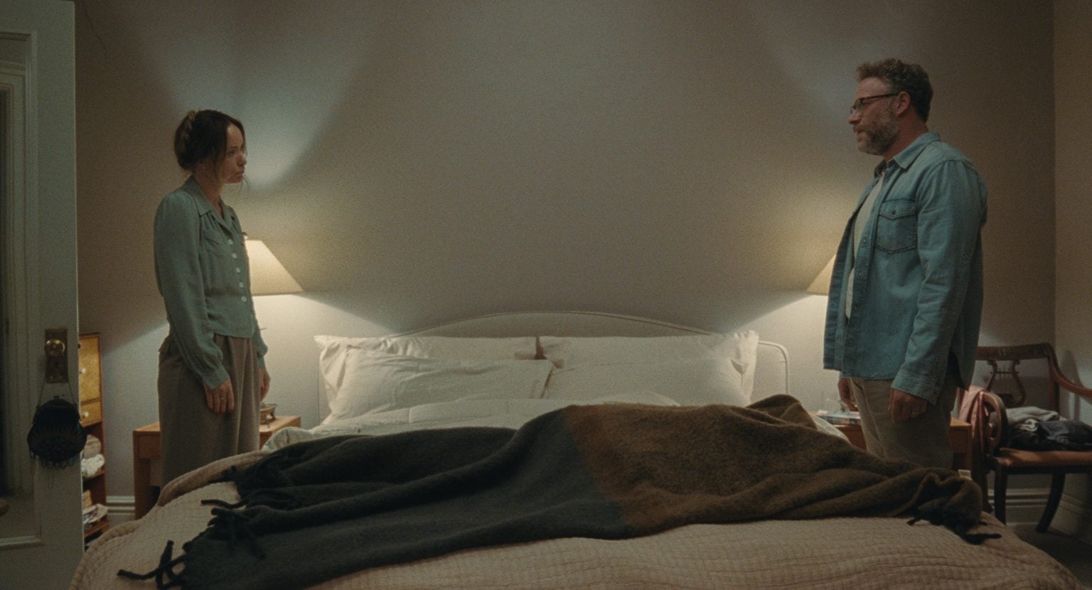 A woman and a man stand on opposite sides of a neatly made bed in a softly lit bedroom, facing each other with neutral expressions.