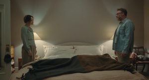 A24 To Release The Hilarious Olivia Wilde & Seth Rogen Comedy ‘The Invite’ On 4K UHD Blu-Ray This November A woman and a man stand on opposite sides of a neatly made bed in a softly lit bedroom, facing each other with neutral expressions.