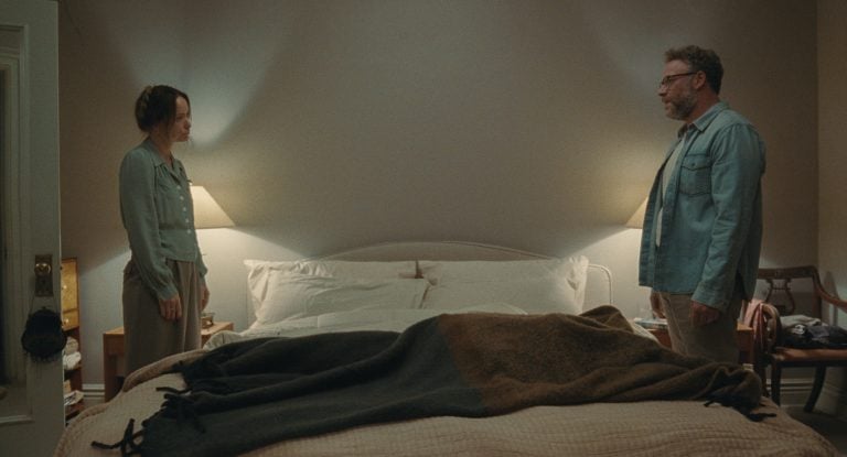 A24 To Release The Hilarious Olivia Wilde & Seth Rogen Comedy ‘The Invite’ On 4K UHD Blu-Ray This November A woman and a man stand on opposite sides of a neatly made bed in a softly lit bedroom, facing each other with neutral expressions.