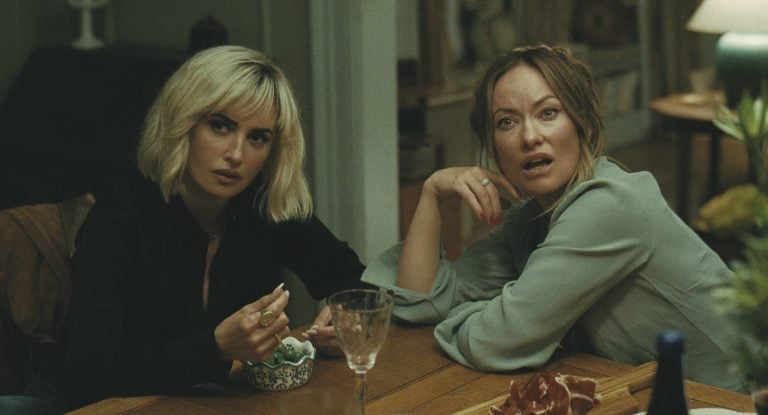 Two women sit at a wooden table with food and drinks, looking intently off-camera with serious and surprised expressions.