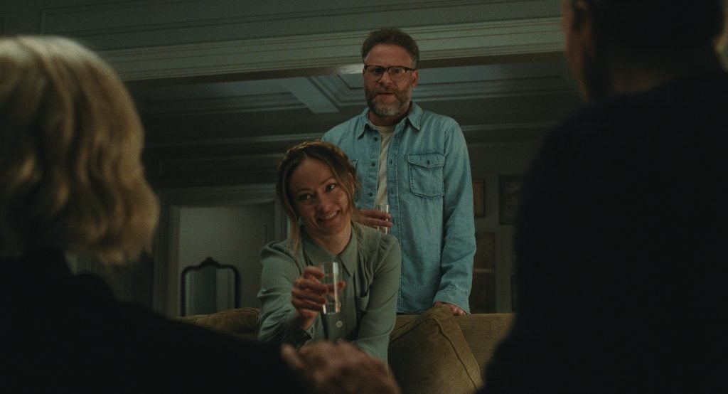 A woman holding a glass of water sits on a couch, smiling at two people in the foreground, while a man stands behind her with a drink in hand.