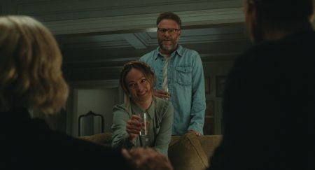 A woman holding a glass of water sits on a couch, smiling at two people in the foreground, while a man stands behind her with a drink in hand.