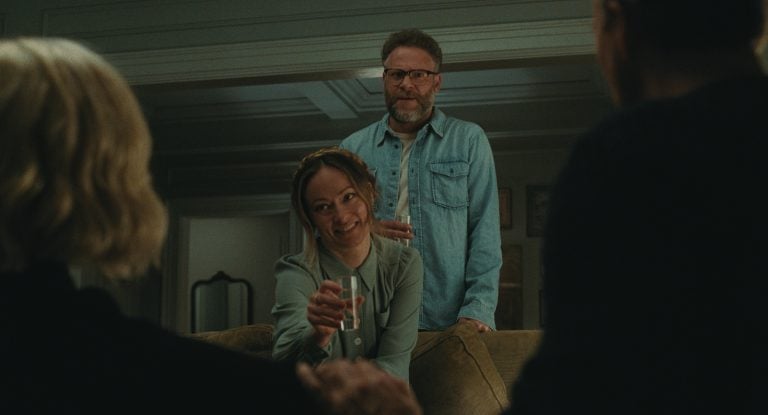 ‘The Invite’ (2026) Review – A Razor-Sharp Dramedy That Is Equally Hilarious And Deeply Insightful About Modern Relationships A woman holding a glass of water sits on a couch, smiling at two people in the foreground, while a man stands behind her with a drink in hand.