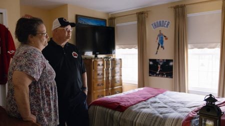 Two people stand in a bedroom decorated with basketball posters, looking toward the windows. A bed, TV, and dresser are visible in the room.