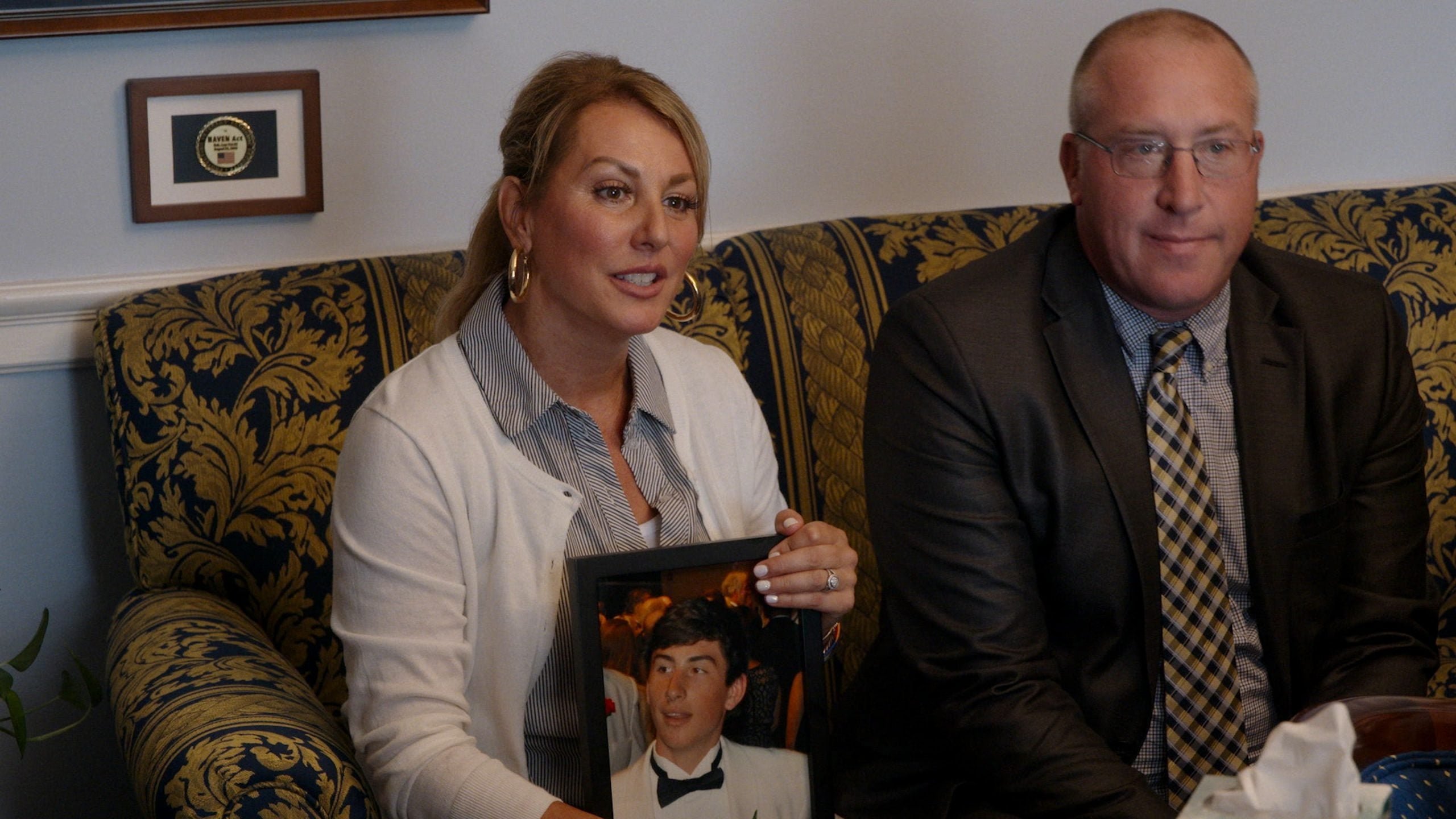 A woman sits on a patterned couch holding a framed photo of a young man in a tuxedo, beside a man in a suit.