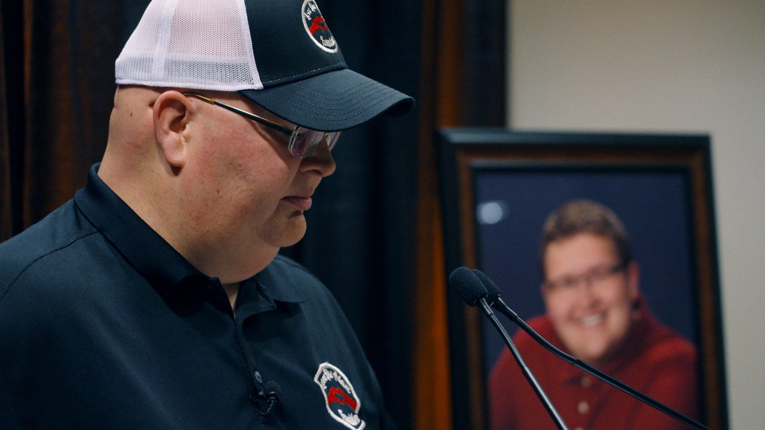 A man in a black polo and cap speaks at a microphone next to a framed photograph of another man in the background.