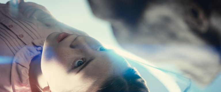 A young girl in a pink shirt looks intently at an indistinct object in front of her, with bright light illuminating her face.