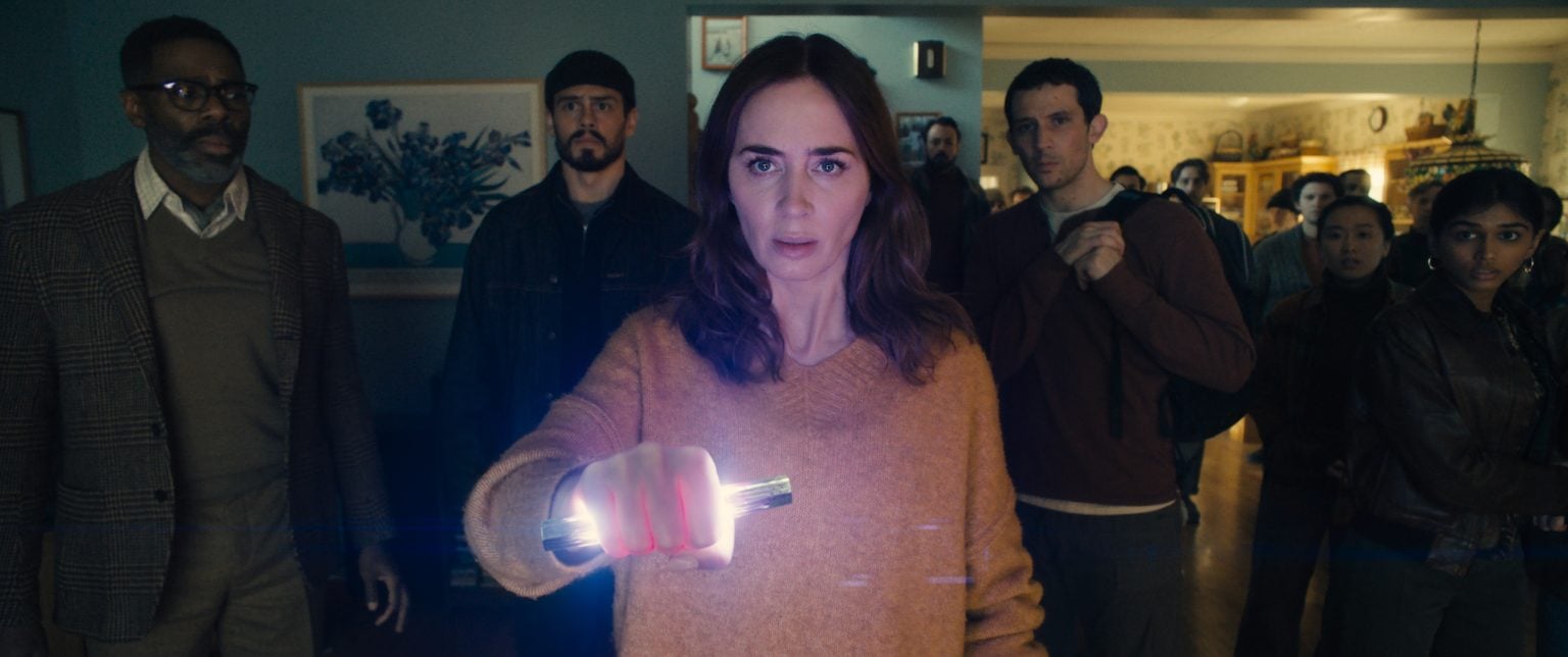 ‘Disclosure Day’ Review – Steven Spielberg Returns To The Blockbuster Space To Remind Us Why He’s One Of Our Best A woman in a beige sweater holds a glowing object, surrounded by a group of people standing in a dimly lit room, all looking ahead with serious expressions.