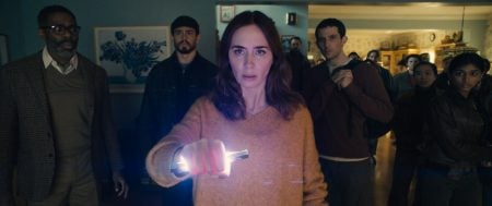 A woman in a beige sweater holds a glowing object, surrounded by a group of people standing in a dimly lit room, all looking ahead with serious expressions.
