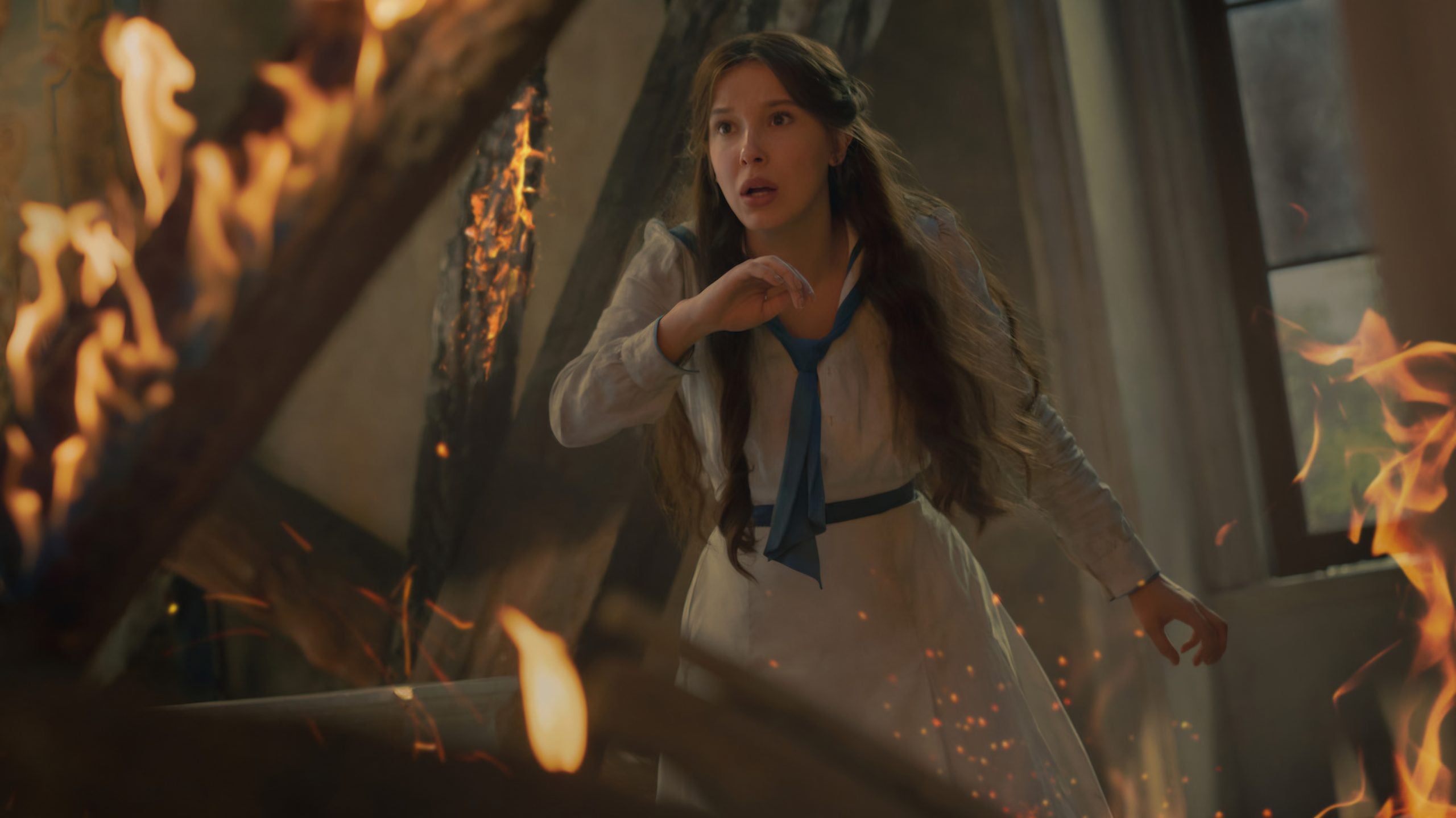 A young woman in a long dress and blue necktie looks concerned as she stands among burning debris inside a building.