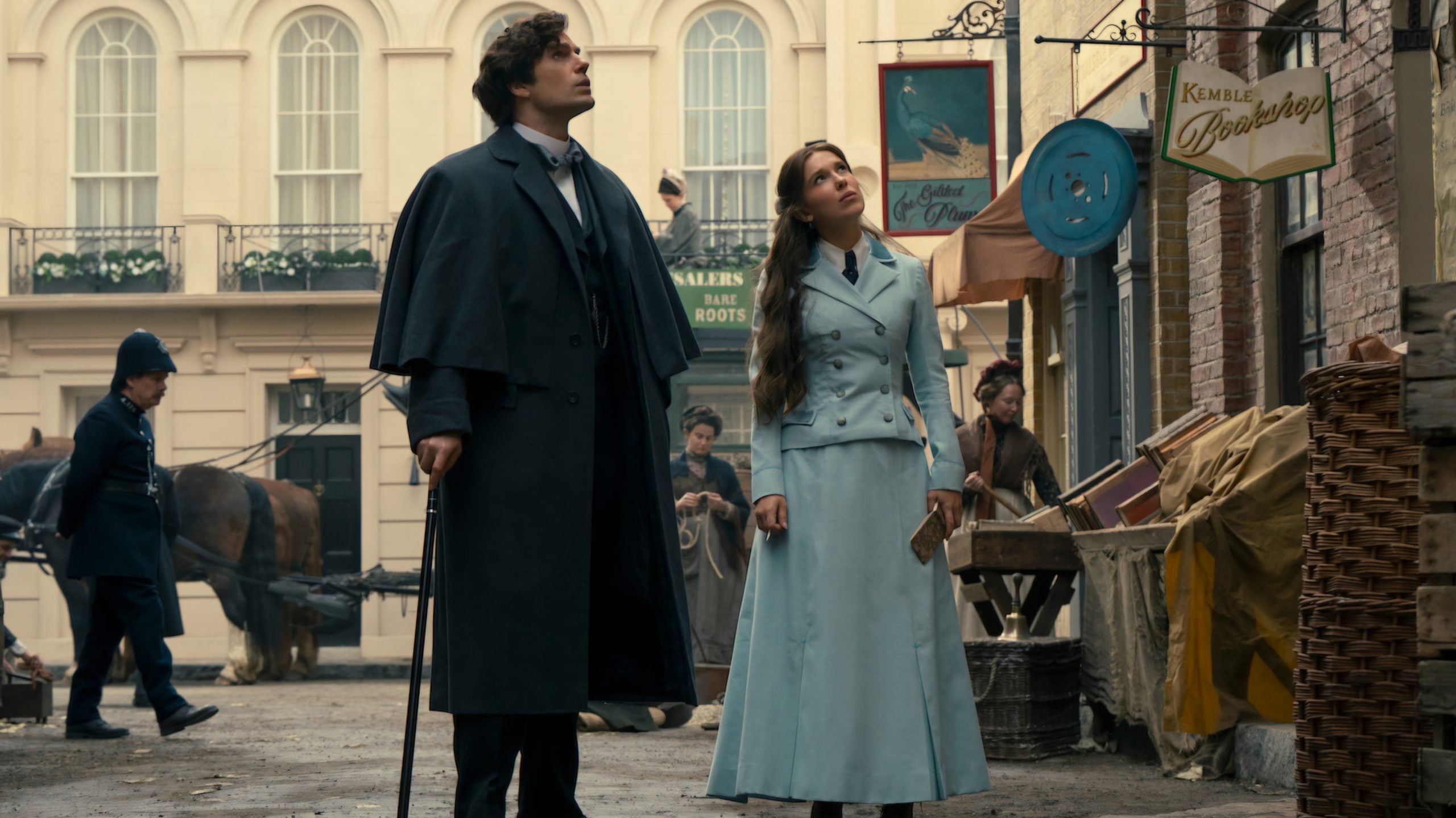 A man with a cane and a woman in light blue Victorian clothing stand on a cobblestone street, looking up, with a shop and other people in the background.