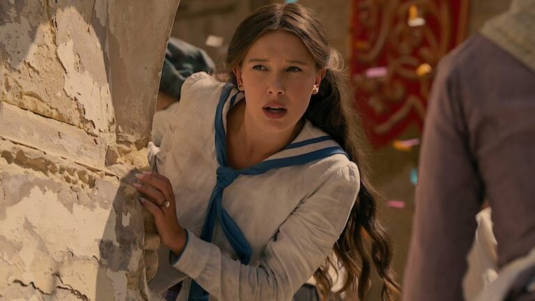 ‘Enola Holmes 3’ Review: Millie Bobby Brown’s Netflix Mystery Finds Charm, Darkness, and a Few Fingerprints of Franchise Fatigue A young woman in a white and blue sailor-style outfit crouches behind a stone wall, looking alert and concerned as colorful confetti falls in the background.