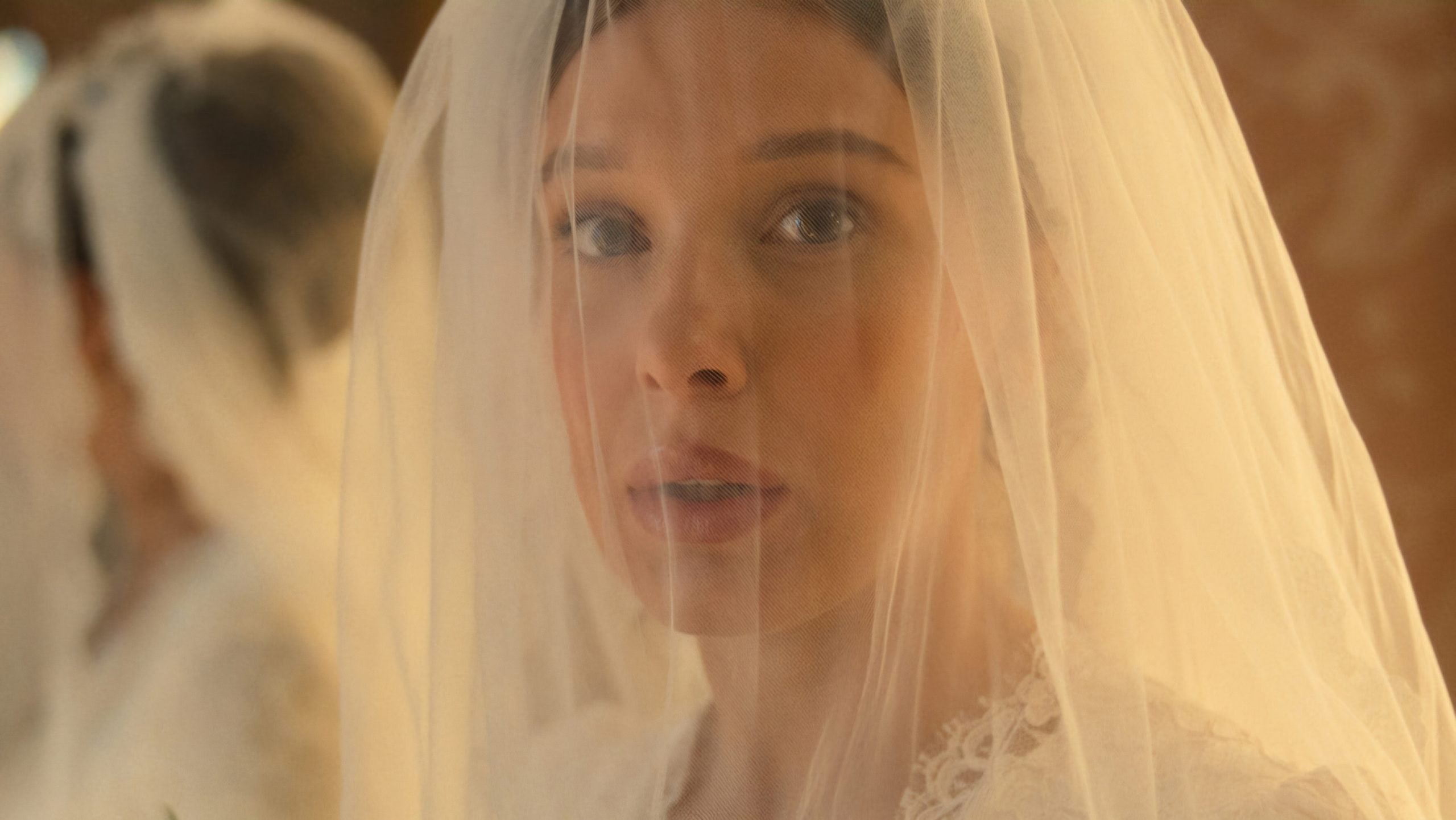 A woman wearing a white lace dress and a veil looks forward with a neutral expression. Another veiled figure is blurred in the background.