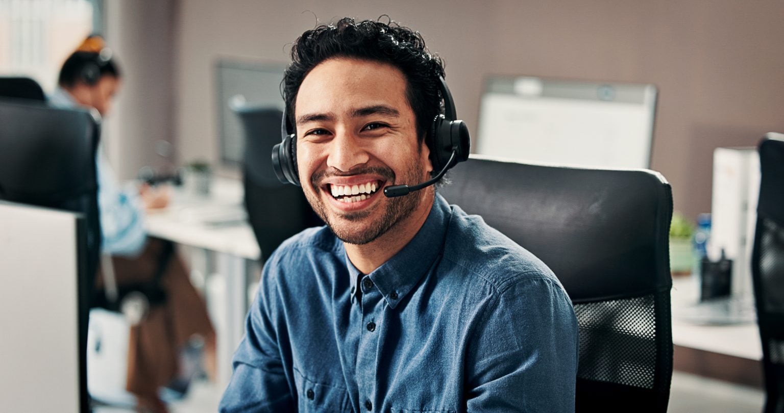 A man wearing a headset and a blue shirt sits at a desk in an office, smiling at the camera.