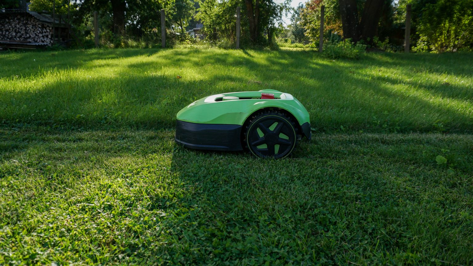 How Do You Choose The Best Electric Lawn Mower? A green robotic lawn mower cuts grass on a sunny lawn with trees and a fence in the background.
