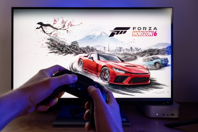 Why Forza Horizon 6’s Wheelspins Drive Hype Person holding a game controller in front of a screen displaying the Forza Horizon 6 video game title and artwork featuring a red sports car and a blue SUV on a racetrack.