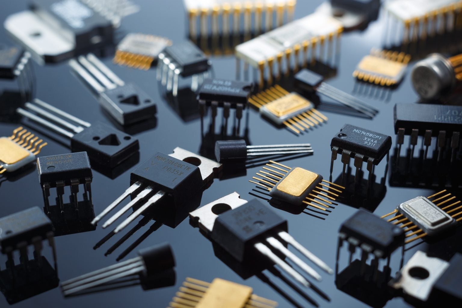A variety of electronic components, including integrated circuits and transistors, are scattered on a reflective black surface.