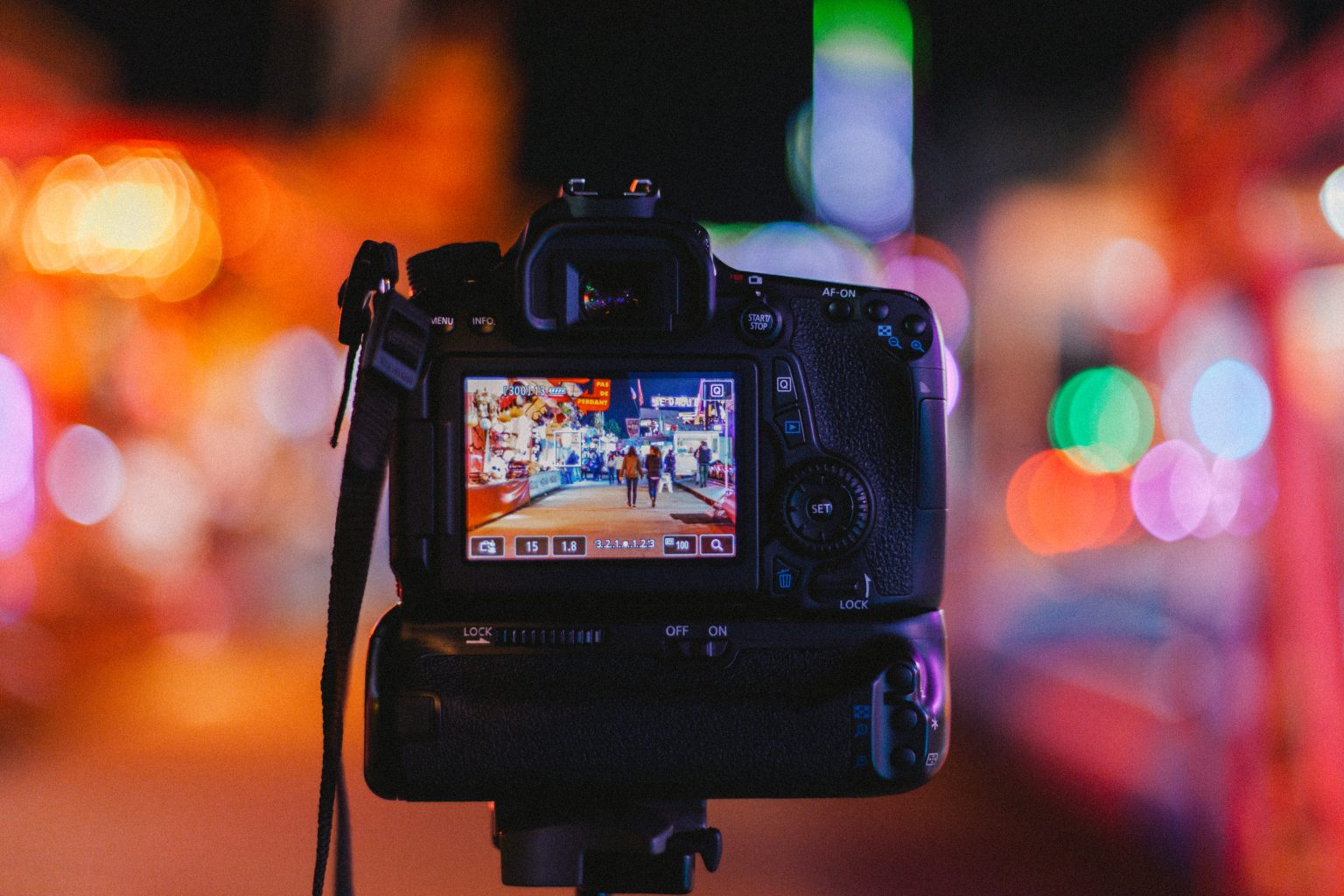 A digital camera on a tripod displays a sharp image of a lively street scene, while the background is blurred with colorful lights.