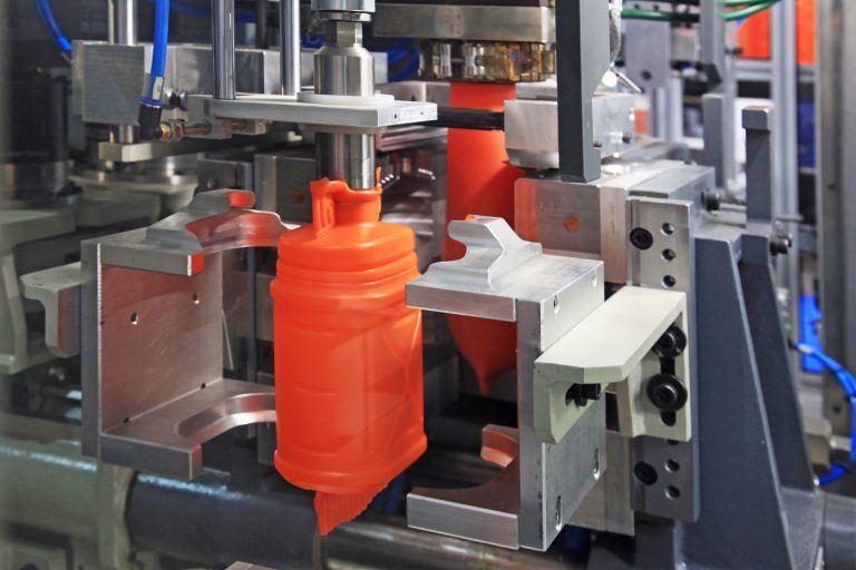 Stop Errors In 2K Plastic Injection Moulding Close-up view of an orange plastic bottle being formed in an industrial blow molding machine.