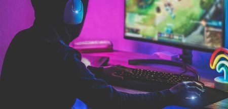 Person wearing headphones sits at a desk playing a video game on a computer monitor, illuminated by pink and blue lighting.