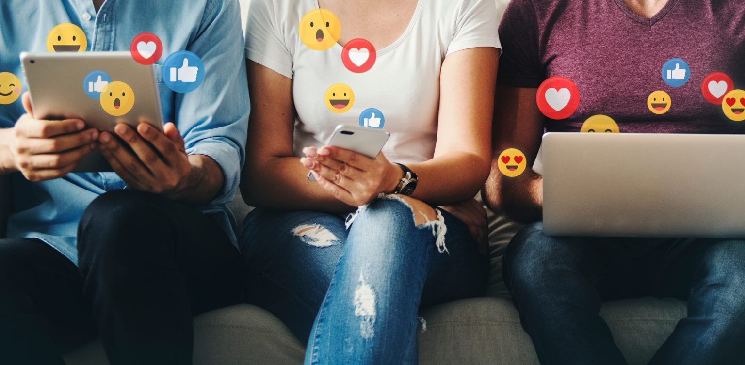 Three people sit side by side using a tablet, smartphone, and laptop, with floating social media reaction icons like thumbs up and hearts.