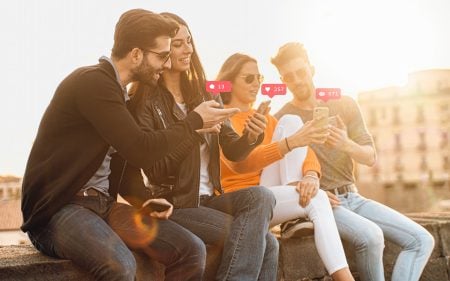 Four young adults sitting outdoors, smiling and using smartphones; social media like and heart notification icons float above their phones.