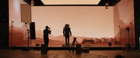 A person in a spacesuit stands on a set with a Mars-like background while a camera operator films; studio lights and equipment are visible.
