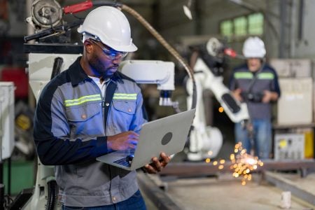 Why Manufacturers Need Better Visibility Across Product Portfolios A worker in safety gear uses a laptop in an industrial setting with robotic machinery and sparks in the background.