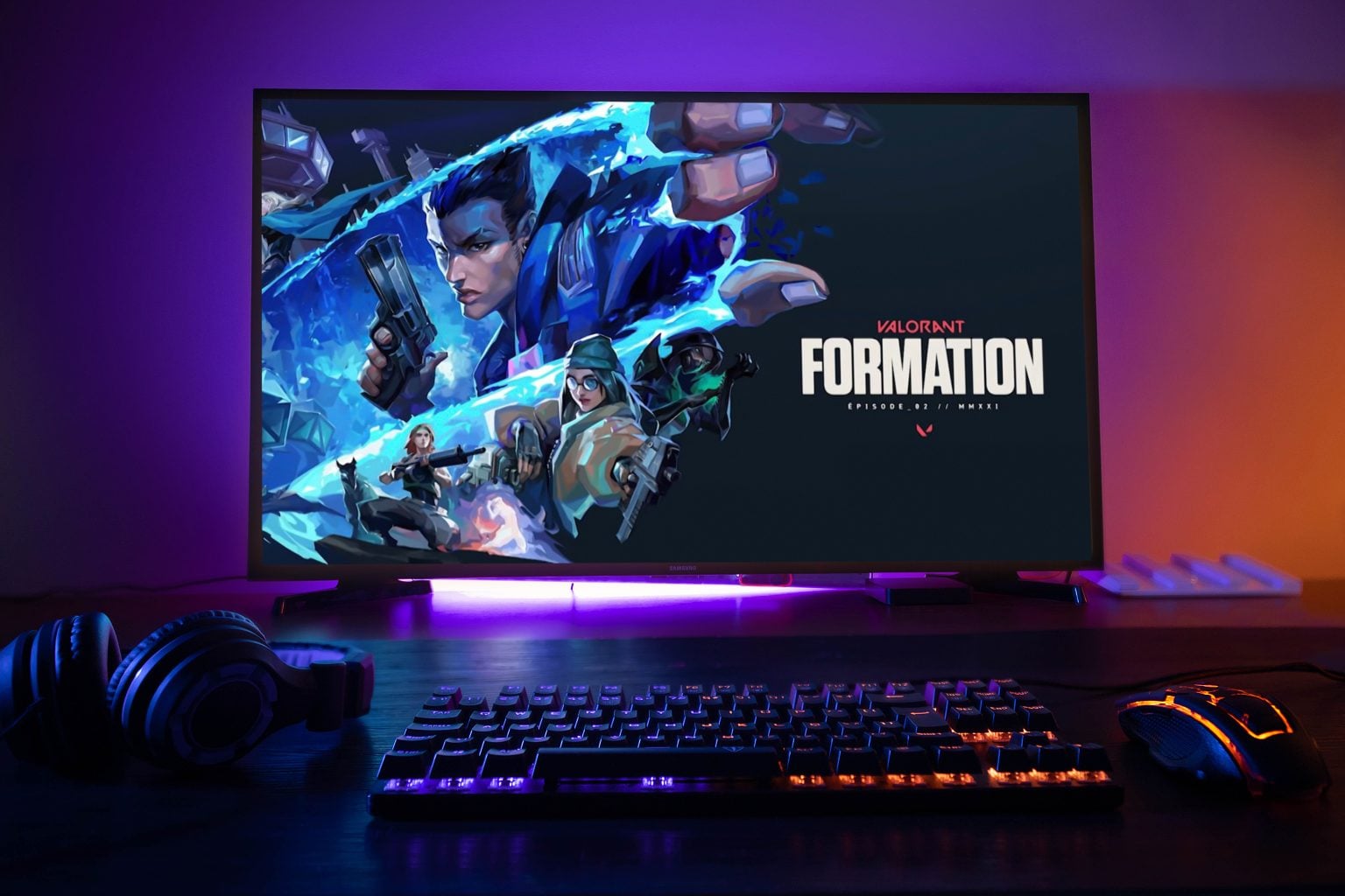 A computer monitor displays the Valorant: Formation game screen, with a keyboard, mouse, and headphones on a desk in a dimly lit room.