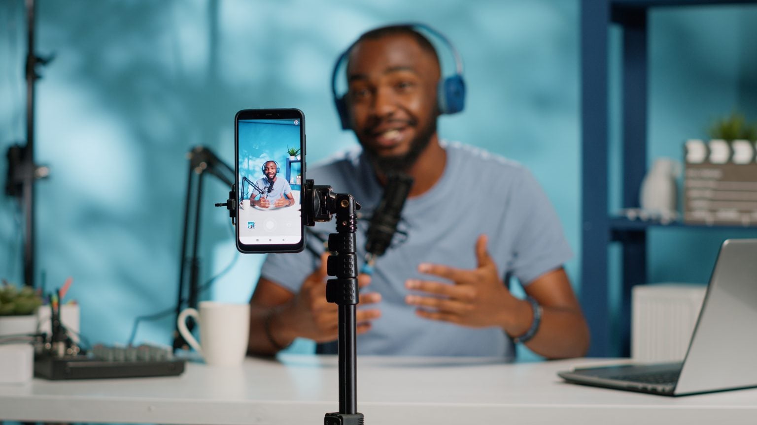 A man wearing headphones speaks into a microphone while recording himself on a smartphone mounted on a tripod at a desk with a laptop and coffee mug.